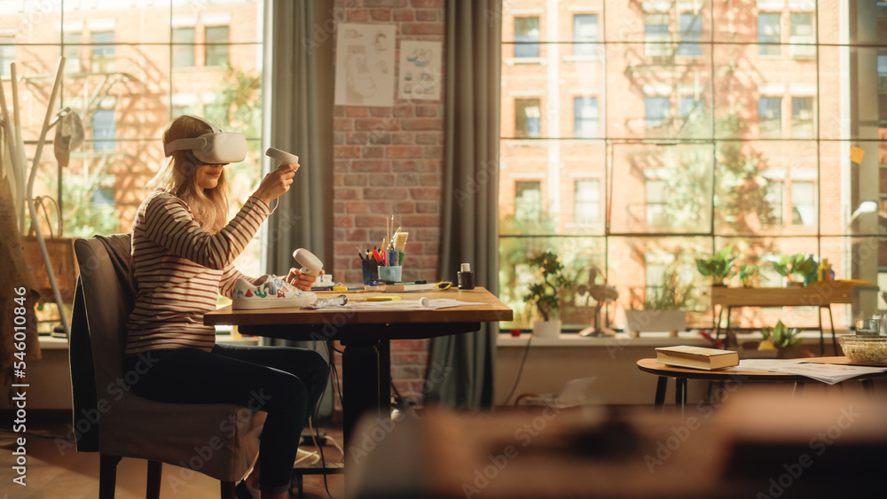 Woman Sitting Behind the Desk at Home or Creative Agency, Using Virtual Reality Headset and Controllers. Young Female Working from Living Room, Experiencing Futuristic Social Media Services. Woman Sitting Behind the Desk at Home or Creative Agency, Using Virtual Reality Headset and Controllers. Young Female Working from Living Room, Experiencing Futuristic Social Media Services.
