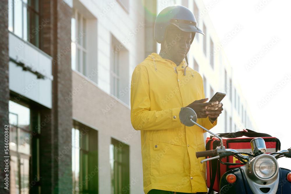 Young courier in safety helmet and yellow jacket standing by bike and looking through addresses of customers in smartphone outdoors
