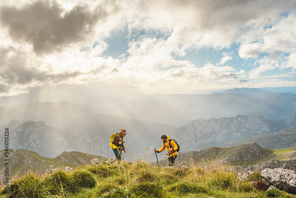 Obraz premium couple of hikers, man and woman, reaching the top of a mountain on a day with clouds and sun. Two mountaineers hiking equipped with backpacks and trekking poles. mountainous landscape from height.