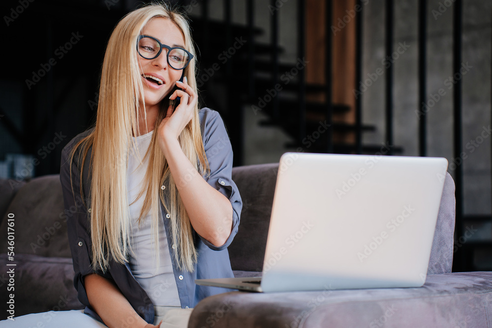 Cheerful young Italian girl in casual sitting on cozy sofa with laptop makes call at home. Creative interior designer speaks with client using cellphone. Businesswoman communicates with partner. Cheerful young Italian girl in casual sitting on cozy sofa with laptop makes call at home. Creative interior designer speaks with client using cellphone. Businesswoman communicates with partner.