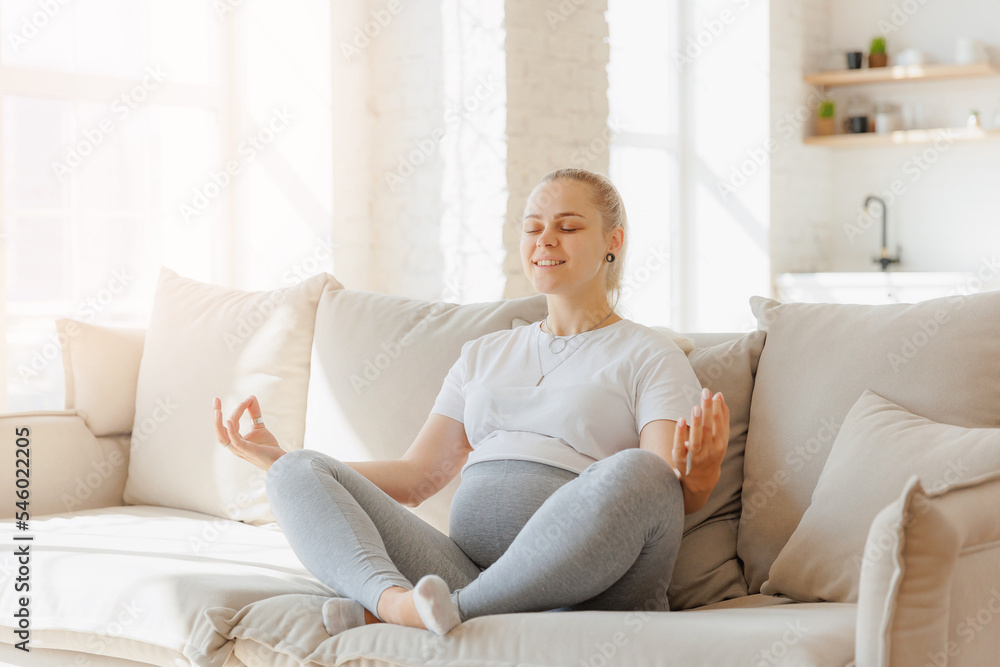 Concept meditating health pregnancy. Portrait beautiful young pregnant woman practicing yoga at home on sofa, background light kitchen Concept meditating health pregnancy. Portrait beautiful young pregnant woman practicing yoga at home on sofa, background light kitchen