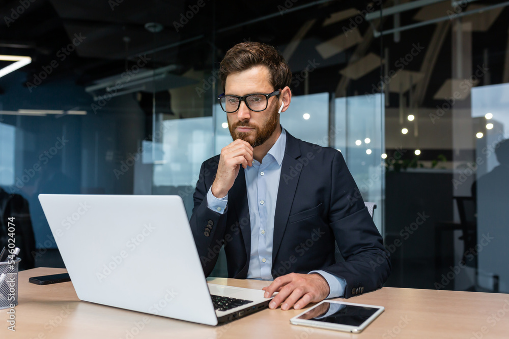 Senior businessman in glasses and beard is thinking about a decision, man is working inside the office and using laptop at work, mature investor in a business suit is sitting at the table Senior businessman in glasses and beard is thinking about a decision, man is working inside the office and using laptop at work, mature investor in a business suit is sitting at the table