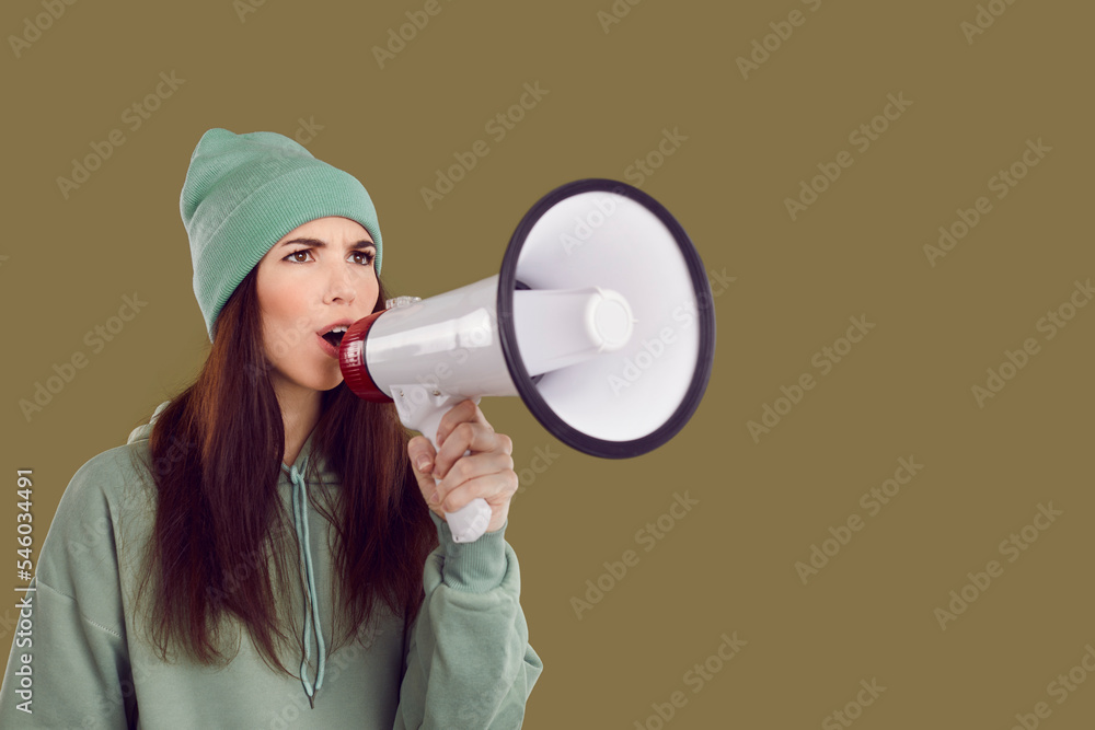 Serious female activist with megaphone in her hand makes loud announcement on brown background. Close up of young woman in casual hoodie and hat with white loudspeaker near copy space. Advertising. Serious female activist with megaphone in her hand makes loud announcement on brown background. Close up of young woman in casual hoodie and hat with white loudspeaker near copy space. Advertising.