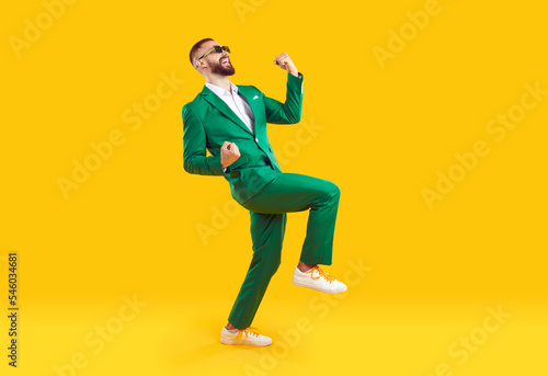 Young man celebrating success. Happy funny joyful excited guy in stylish green party suit and cool glasses raising fist up and dancing isolated on bright yellow background. Full length shot, side view photo