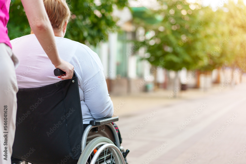 Obraz premium Back view of young woman helping mature woman in wheelchair in the city