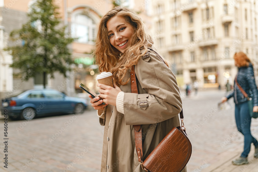 Young woman with curly blonde hair using the phone with a cup of coffee in hands on the city streets