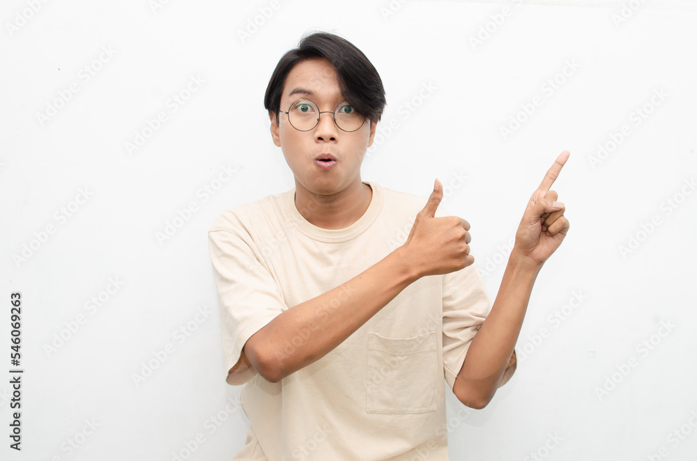 asian young man in casual beige tshirt pointing finger showing something good. man with glasses shocked happily pointing up with copy space isolated over white background. advertisment model concept. asian young man in casual beige tshirt pointing finger showing something good. man with glasses shocked happily pointing up with copy space isolated over white background. advertisment model concept.