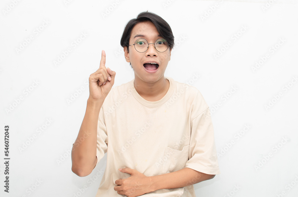 asian young man in casual beige tshirt pointing finger showing something good. man with glasses shocked happily pointing up with copy space isolated over white background. advertisment model concept. asian young man in casual beige tshirt pointing finger showing something good. man with glasses shocked happily pointing up with copy space isolated over white background. advertisment model concept.
