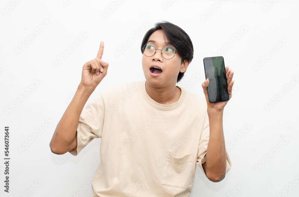 excited young asian man in beige tshirt holding and showing phone screen with happy euphoric shocked wow expression. man pointing to screen phone with thumb up. billboard model advertisment concept. excited young asian man in beige tshirt holding and showing phone screen with happy euphoric shocked wow expression. man pointing to screen phone with thumb up. billboard model advertisment concept.