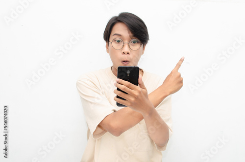excited young asian man in beige tshirt holding and showing phone screen with happy euphoric shocked wow expression. man pointing to screen phone with thumb up. billboard model advertisment concept.