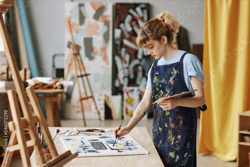 Young inspired woman in apron painting new masterpiece on canvas with acrylic paints while holding palette with mixed colors during work