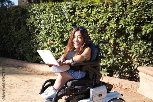 фотографія young disabled woman in wheelchair and reduced mobility with her laptop computer teleworking in the park