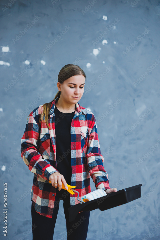 A beautiful young woman in a checkered shirt is renovating her room, holding a paint roller