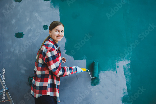 Young smiling pretty woman in shirt holding roller in right hand and painting wall. Repair in the room.