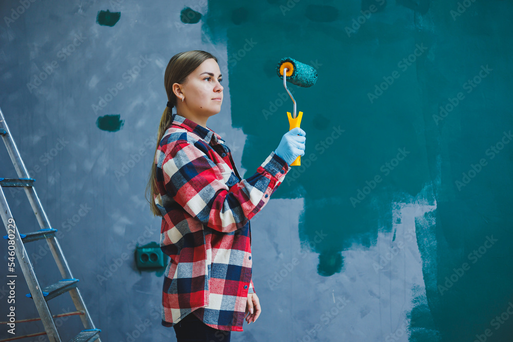 Fototapeta premium A young beautiful woman designer makes repairs, paints a gray wall with green paint with a roller. Plaid shirt, long hair and jeans.