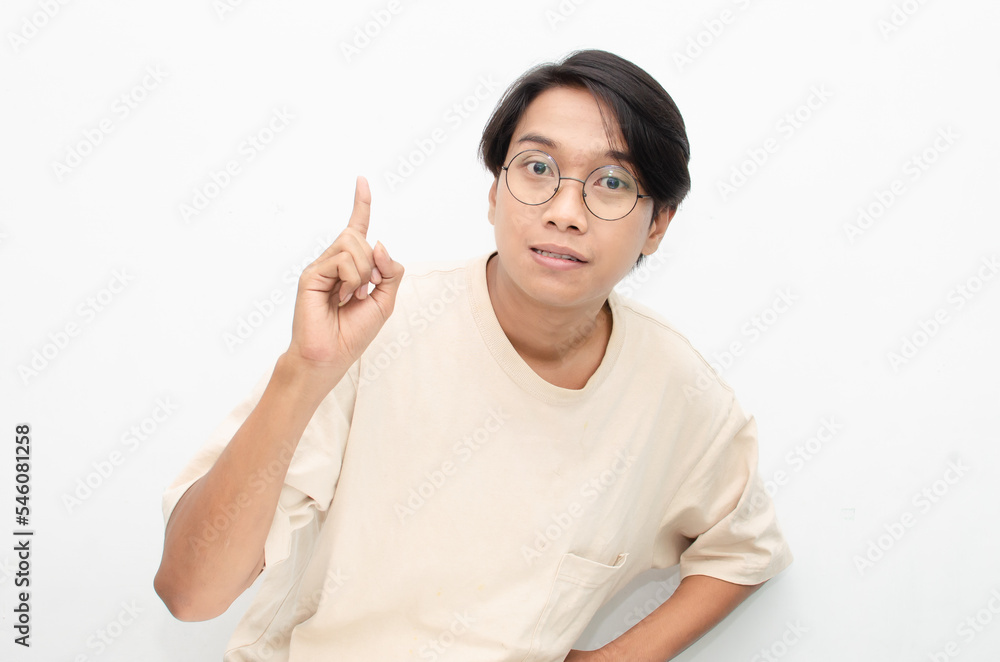 asian young man in casual beige tshirt pointing finger showing something good. man with glasses shocked happily pointing up with copy space isolated over white background. advertisment model concept. asian young man in casual beige tshirt pointing finger showing something good. man with glasses shocked happily pointing up with copy space isolated over white background. advertisment model concept.