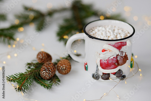 Winter table composition. Metal white cup with Santa Claus with cocoa and marshmallows, decorated with pine cones and branches and garland on a white table