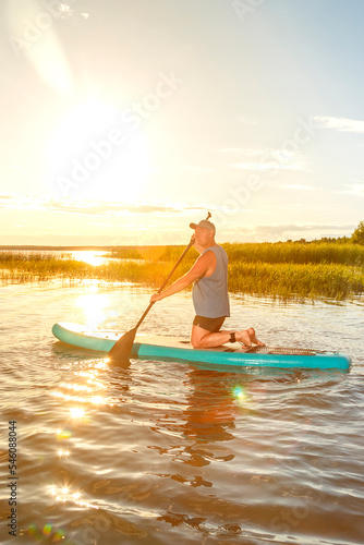 a man in shorts and a T-shirt on a SUP board with an oar floats on the water against the background of the setting sun.