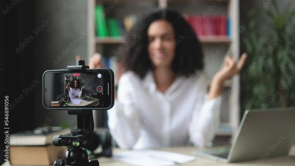 Blurred relaxed young woman in casual wear sitting on workplace with modern laptop and meditating with closed eyes during recording video blog. Focus on modern smartphone fixed on tripod. Blurred relaxed young woman in casual wear sitting on workplace with modern laptop and meditating with closed eyes during recording video blog. Focus on modern smartphone fixed on tripod.