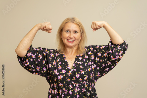 Caucasian blonde woman in a dark patterned dress raising her arms and flexing her biceps muscle isolated over beige background. Strong and brave modern woman concept.