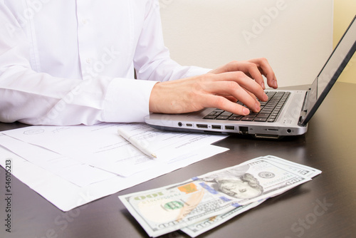 close-up of human hands typing on a computer keyboard. in the foreground are dollar bills