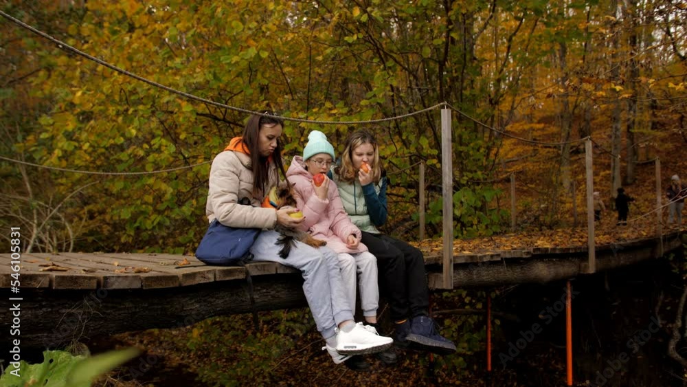 A woman in an embrace with her daughters are sitting together on a wooden bridge across the river in the autumn forest and eating apples. A woman in an embrace with her daughters are sitting together on a wooden bridge across the river in the autumn forest and eating apples.