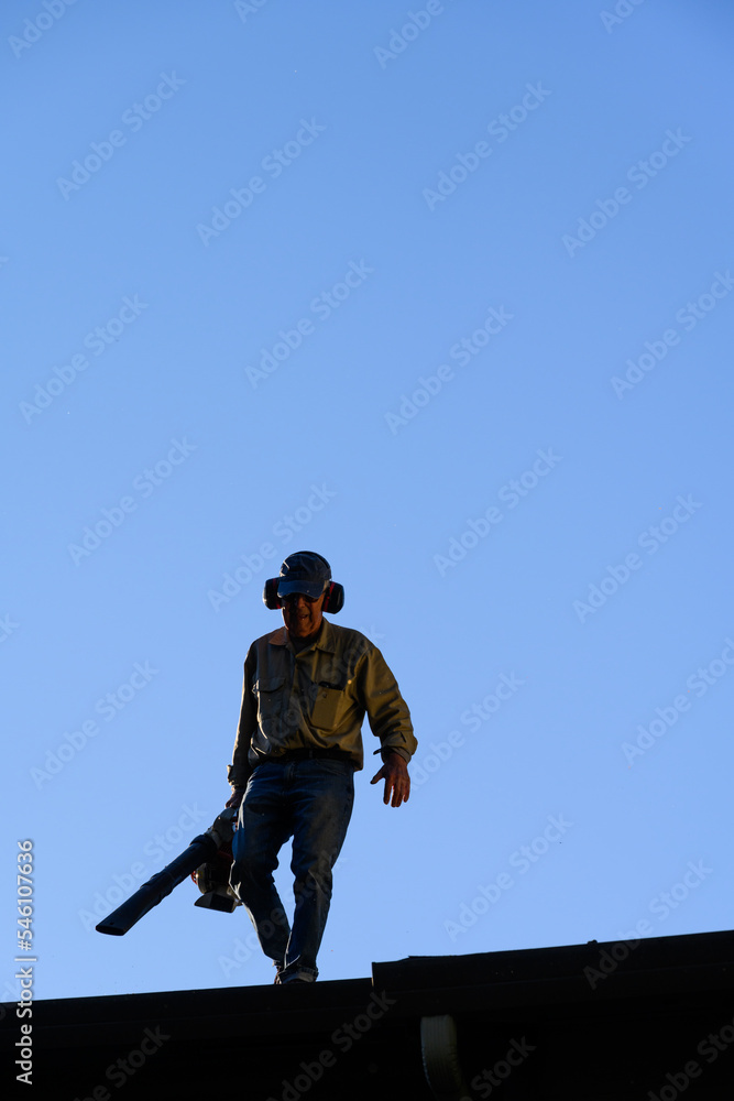 Silhouette of senior man with gas powered leaf blower cleaning roof gutters on an apartment building, fall maintenance
