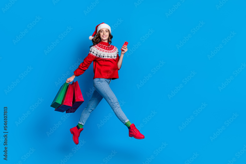 Full length photo of funky positive girl dressed red pullover jumping holding x-mas bargains device empty space isolated blue color background Full length photo of funky positive girl dressed red pullover jumping holding x-mas bargains device empty space isolated blue color background