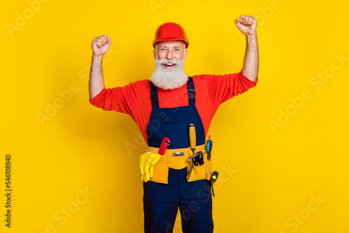 Photo of positive strong old guy dressed uniform overall red hardhat rising hands showing biceps isolated yellow color background © deagreez