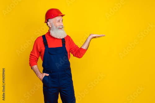 Photo of positive funky senior guy dressed uniform overall red hardhat looking holding arm empty space isolated yellow color background