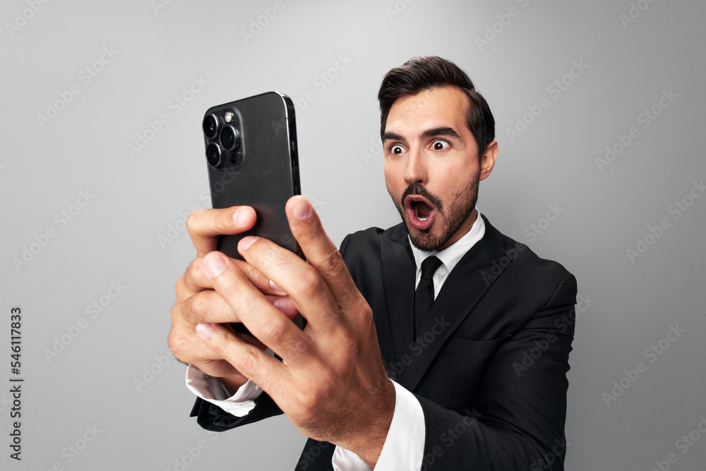 Man businessman holding phone in hand surprised looking at screen video call and selfies and smile with teeth. Close-up wide angle photo gray isolated background Man businessman holding phone in hand surprised looking at screen video call and selfies and smile with teeth. Close-up wide angle photo gray isolated background