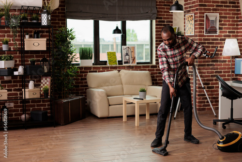 Single African American man doing house cleaning, concentrating while vacuuming the floor. Doing housework before sitting down to work on his computer from home. Freelance worker.