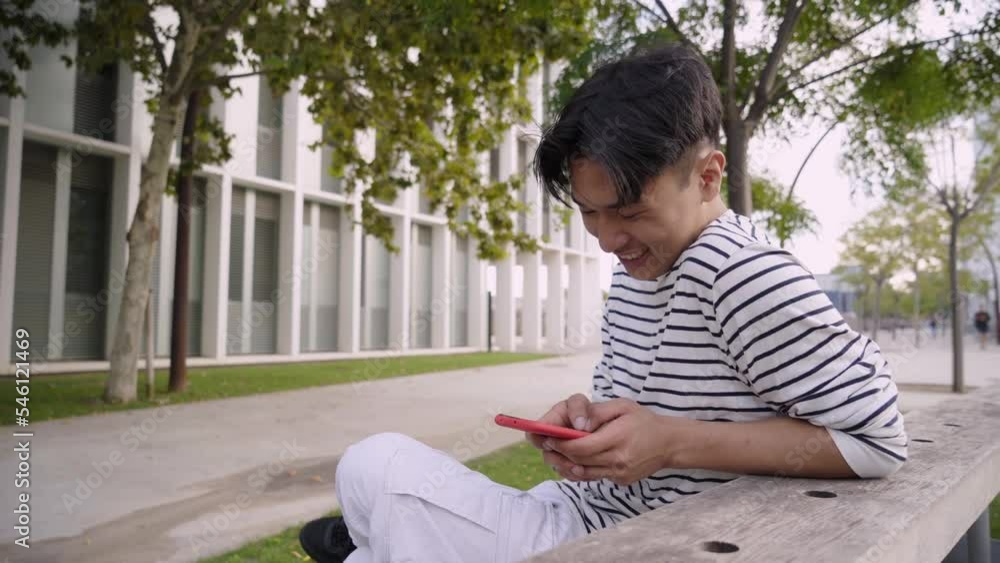 Funny Asian guy at university campus sitting on a bench chatting with a smart phone. Student using mobile cell and laughing outdoors. Cheerful people socializing on social media millennial generation Funny Asian guy at university campus sitting on a bench chatting with a smart phone. Student using mobile cell and laughing outdoors. Cheerful people socializing on social media millennial generation