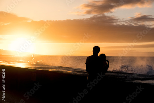 Backlight of a father and his little toddler son watching a sunset in front of the sea in Tenerife, Canary Islands