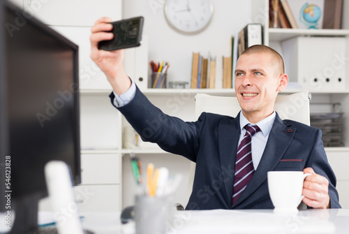 Business consultant male working in an office takes a selfie while sitting at the workplace with a cup of coffee