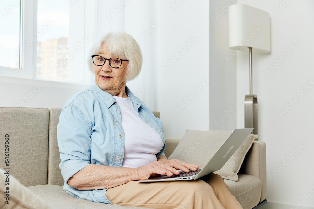 a joyful old lady is sitting on a beige sofa stylishly dressed and working from home in a comfortable environment holding a laptop on her lap a joyful old lady is sitting on a beige sofa stylishly dressed and working from home in a comfortable environment holding a laptop on her lap