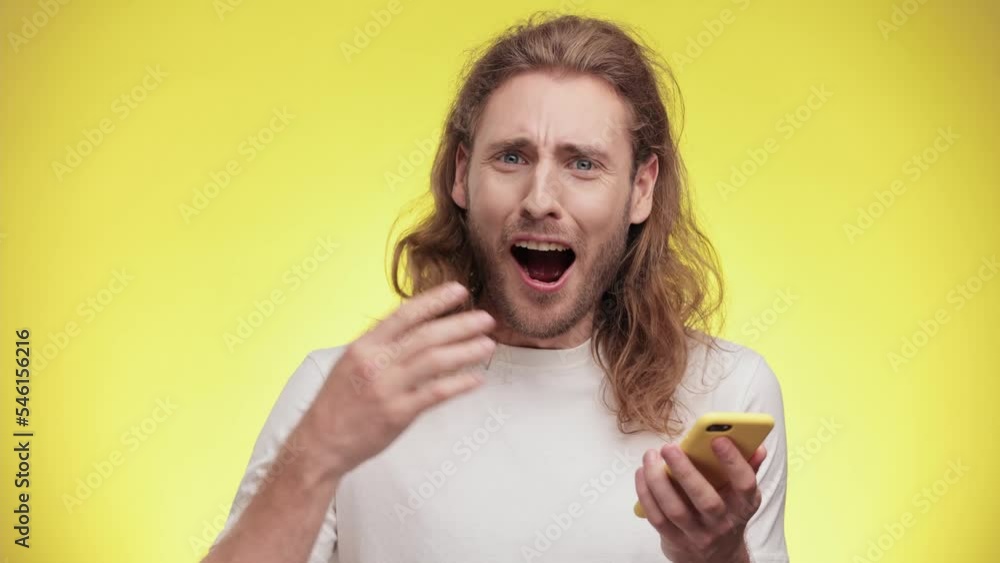 Young Caucasian man wears white T-shirt hold use mobile cell phone typing browsing chatting send sms doing online shopping order booking tour isolated on yellow wall background studio portrait. Young Caucasian man wears white T-shirt hold use mobile cell phone typing browsing chatting send sms doing online shopping order booking tour isolated on yellow wall background studio portrait.