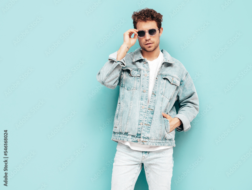 Portrait of handsome confident stylish hipster lambersexual model with curly hairstyle. Sexy man dressed in jeans jacket. Fashion male isolated on blue wall in studio. In sunglasses Portrait of handsome confident stylish hipster lambersexual model with curly hairstyle. Sexy man dressed in jeans jacket. Fashion male isolated on blue wall in studio. In sunglasses