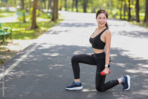 Strong and beautiful asian fitness female sportswoman lifting dumbbell weight training after finish morning run outdoor. Happy smiling athlete woman doing squats exercise under trees in the park