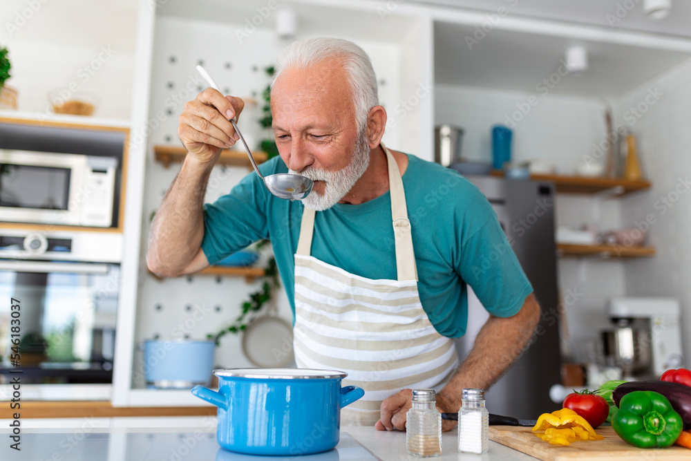 Happy retired senior man cooking in kitchen. Retirement, hobby people concept. Portrait of smiling senior man holding spoon to taste food Happy retired senior man cooking in kitchen. Retirement, hobby people concept. Portrait of smiling senior man holding spoon to taste food