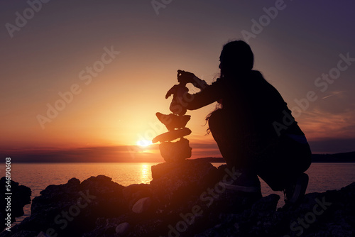 Silhouette of a woman balancing rocks and stones on the ocean sea coast at sunset sunrise time. © astrosystem