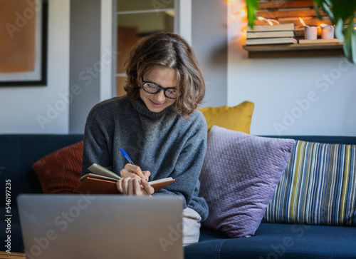 Young woman in a warm sweater sitting on a sofa with a laptop making notes in a notebook, work and education online concept © olezzo