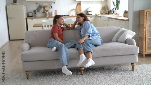 Young Caucasian woman gossips with daughter sits on couch and asks about school friends or favorite hobbies. Teenage girl in casual clothes shares secrets with mother or beg permission to go to party