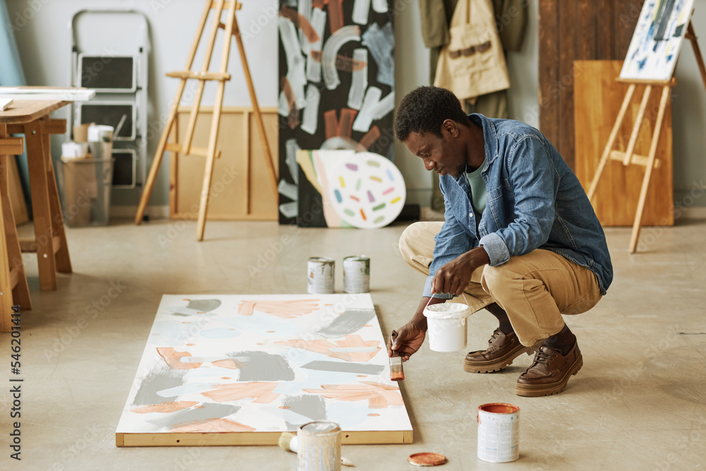 Young black man sitting on squats and bending over canvas with unfinished artwork while painting it in studio or classroom - obrazy, fototapety, plakaty 