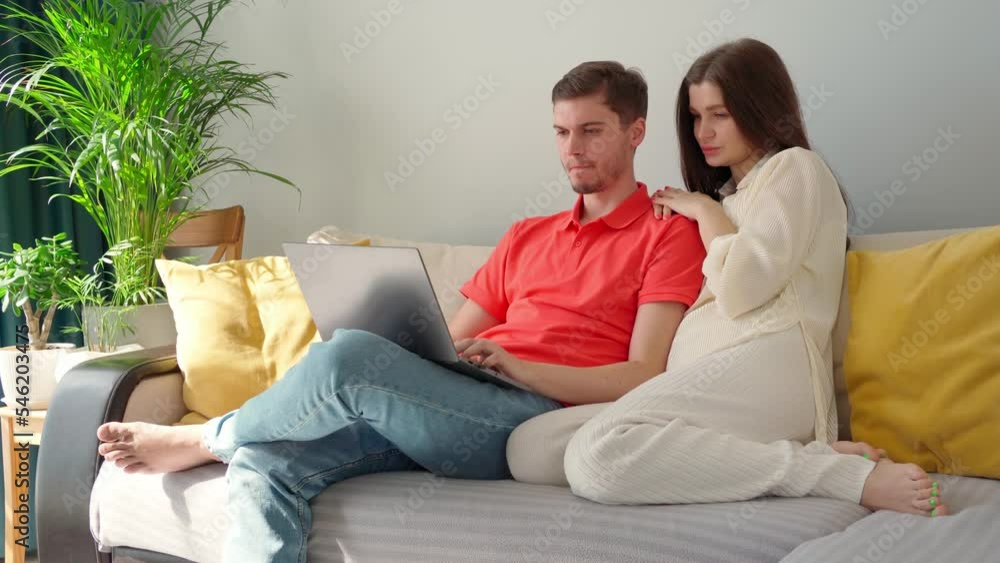 young woman and man on the sofa in the living room looking at something in the laptop. a man is typing in a laptop, a woman behind him is looking at the laptop with interest