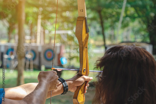 Hands of young man aims archery bow and arrow to colorful target in shooting range during training. Exercise and concentration with outdoor archery. Selective focus on hand. Sport, Recreation concept.