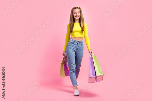 Full length photo of pretty sweet school girl dressed yellow crop top walking holding bargains isolated pink color background