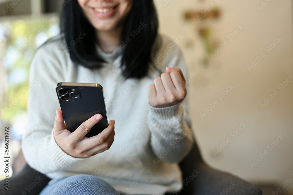 Fototapeta premium Happy young Asian woman using her smartphone and showing clenched fist. cropped