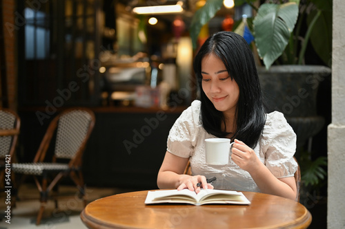 Asian female reading a book and sipping coffee while relaxing in the beautiful coffee shop.