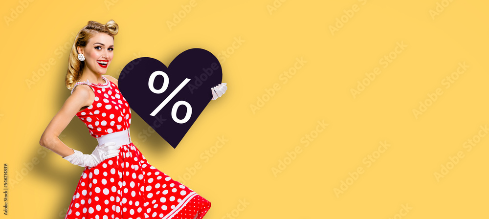 Fototapeta premium Woman holding red paper heart shape with % sign. Portrait image of happy pin up girl. Blond model at retro fashion rockabilly sales ad concept. Black Friday sale. Yellow background.